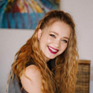 A person with long, curly blonde hair and bright lipstick is smiling while looking at the camera. The background features a colorful abstract painting and a woven basket.